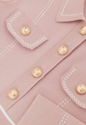 Material tricot roz deschis cu detalii albe cusute. Prezintă două nasturi aurii și buzunare cu clapetă. Textură netedă, elemente de design croite pe măsură.