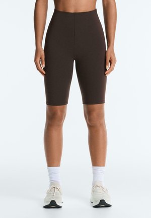 Sötétbarna magas derekú biciklis rövidnadrágot, fehér zoknit és fehér sportcipőt viselő személy karjait lazán oldalt tartva áll.