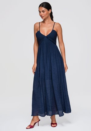 Femme portant une robe longue bleu marine sans manches avec de fines bretelles et des talons hauts rouges ouverts à l'avant, debout devant un fond uni.