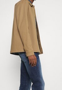 Giacca marrone chiaro con bottoni e colletto, realizzata in cotone testurizzato. Indossata sopra una camicia a quadri blu, abbinata a jeans in denim scuro.