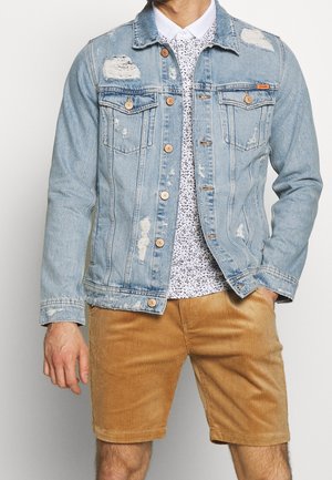 Homme portant une veste en jean bleu clair usée, une chemise blanche à motifs et un short en velours côtelé beige, debout avec une main dans la poche.