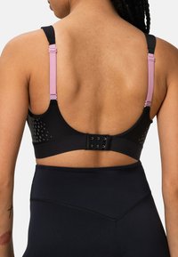 Schwarzer Sport-BH mit durchlöchertem Design, verstellbaren pinken Trägern und einer dreifachen Hakenverschluss auf der Rückseite. Kombiniert mit hochgeschnittenen schwarzen Leggings.
