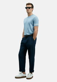 Lichtblauw kortarmshirt met witte accenten, donkere marineblauwe broek en witte sneakers met een gumkleurige zool, gedragen met zwarte zonnebril.