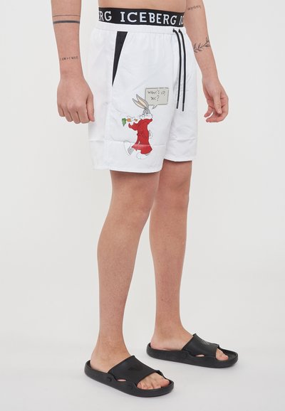 Uomo che indossa pantaloni corti bianchi con un grafiche di Bugs Bunny e il testo "Che succede, dottore?", sandali neri, tatuaggi sugli avambracci, in piedi contro uno sfondo semplice.