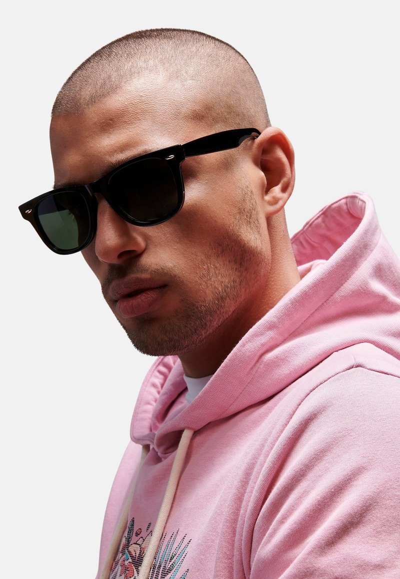 Gafas de sol negras con lentes oscuros y montura cuadrada, llevadas por una persona con una sudadera rosa clara con un estampado gráfico. Acabado suave y mate.