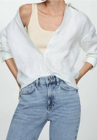 Camisa de lino blanca, abotonada en la parte delantera, con mangas enrolladas sobre una camiseta de tirantes de canalé color beige claro. Combinada con unos pantalones vaqueros de cintura alta, de color azul claro.