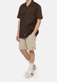 Muž oblečený v hnědé košili s krátkým rukávem na knoflíky, béžových šortkách, bílých ponožkách a bílých teniskách s hnědým logem Nike.