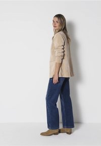 Beige, měkká látková bunda s volným střihem, bočními kapsami a ohrnutými rukávy. Přiřazena k širokým modrým džínům a hnědým botám.