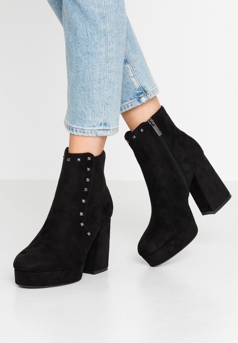 Bottes courtes en daim noir avec un talon haut épais, ornées de clous métalliques sur le côté et une fermeture éclair pour un enfilage facile.