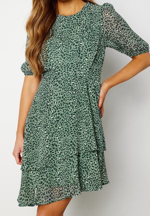 Mujer que lleva un vestido verde de manga corta con falda de capas y estampado de leopardo negro, posando con una mano en la cadera.