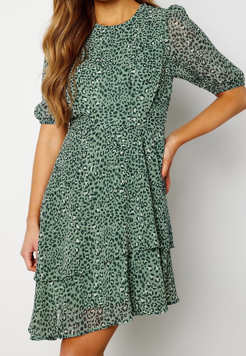 Femme portant une robe verte à manches courtes avec une jupe à volants et un imprimé léopard noir, posant avec une main sur la hanche.