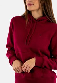 Sudadera con capucha burdeos hecha de tela suave, con un bolsillo tipo canguro en la parte delantera, cordones ajustables y un pequeño logotipo en el pecho.