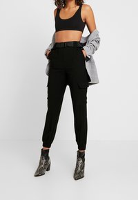 Zwarte cargobroek met elastische boorden en een ceintuur in de taille, gecombineerd met een aansluitende zwarte cropped top en een grijs geruit blazer.