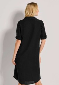 Schwarzes Hemdkleid aus leichtem Stoff, mit Kragen und aufgerollten Ärmeln, in lockerer Passform und seitlichem Schlitz für Bewegungsfreiheit.