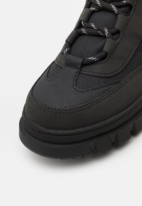Czarne buty trekkingowe z chropowatą cholewką, wykonaną z połączenia gładkich i wzorzystych materiałów, grubą czarną gumową podeszwą oraz kontrastującymi sznurówkami.