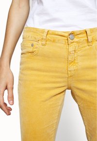 Pantalones de mezclilla amarillos con un corte clásico, que cuentan con un cierre frontal de botón, diseño de cinco bolsillos y un sutil desgastado en la textura de la tela.