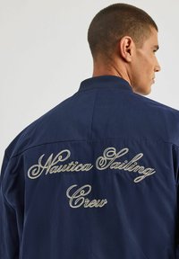 Granatowa kurtka z prążkowanym kołnierzem, z białym haftowanym napisem "Nautica Sailing Crew" na plecach. Gładka tekstura materiału.