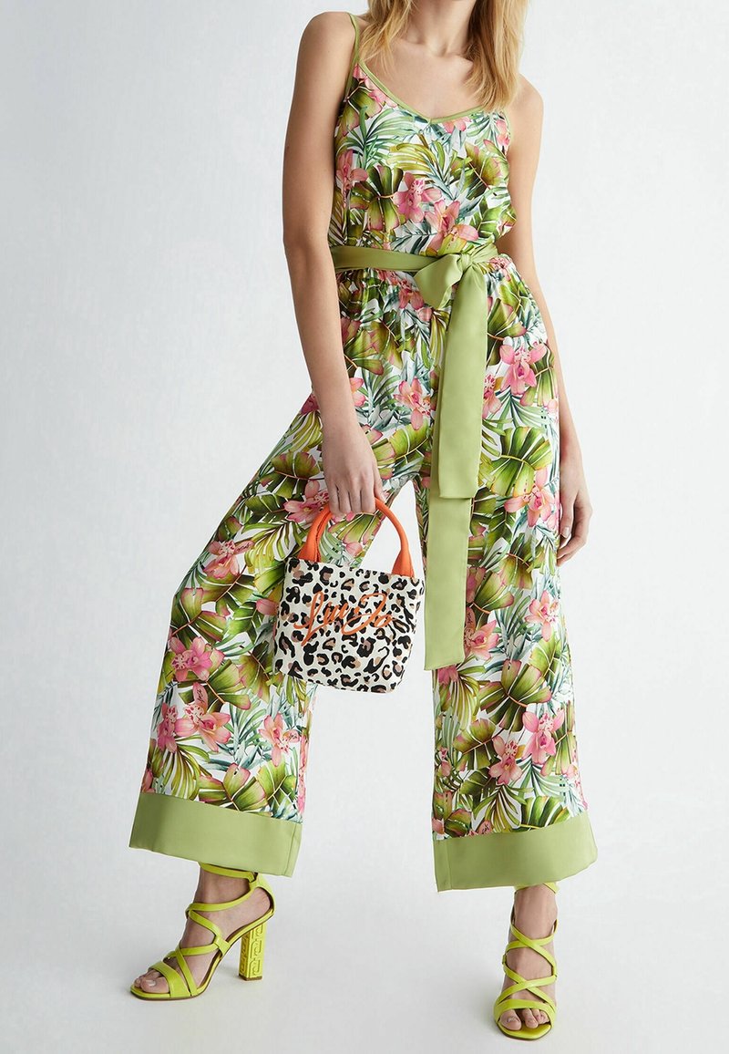 Femme portant une combinaison large à motif floral avec une ceinture verte, tenant un sac à main imprimé léopard avec des poignées oranges, et des talons verts citron à brides.
