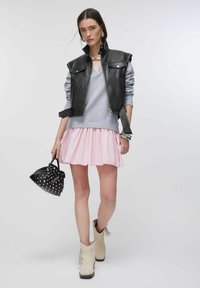 Gilet court en cuir noir porté sur un pull gris, associé à une jupe plissée rose. Bottines beiges à franges et un sac à main noir clouté.