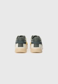 Sneakers met een groene stoffen bovenkant, crème accenten en een gestructureerde beige rubberen zool; heeft een gele detail en donkere banden.