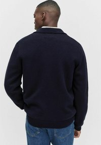 Man som bär en mörk marinblå stickad tröja med krage, vänd bort, tillsammans med blå jeans mot en enkel ljus bakgrund.