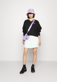 Melns oversized džemperis ar izšūtu logo, balta plisēta svārki, violeta ziedu printa spainīša cepure, violeta plecu soma un melni sporta apavi.