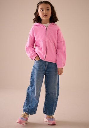 Jeune fille portant un sweat à capuche zippé rose, un jean large bicolore et des baskets roses, debout avec une main dans la poche.