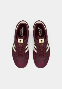 Bordó tenisky s texturovaným svrškem, krémovými pruhy a zlatým logem. Mají tkaničky a gumovou podrážku. Navrženy s kontrastními vzory.