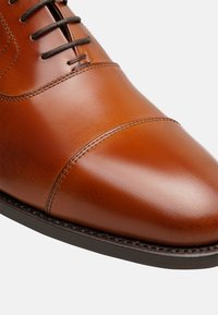 Chaussure habillée en cuir marron avec un design élégant, une texture brillante, un détail de bout cap and des lacets foncés, sur une semelle noire contrastante.