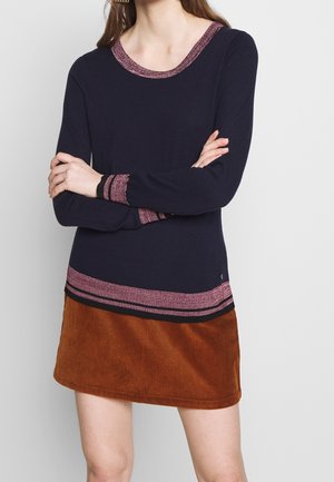 Kobieta ubrana w granatowy sweter z różowo-czarną paskowaną lamówką oraz brązową mini spódnicę sztruksową, ze skrzyżowanymi ramionami.