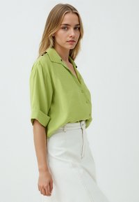 Camicia verde lime a maniche corte con colletto, design abbottonato, abbinata a una gonna bianca a vita alta con dettagli di cucitura visibili.