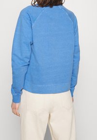 Suéter azul de algodón con mangas raglán, puños y dobladillo acanalados, textura suave y detalle sutil de costura en el escote, combinado con pantalones beige.