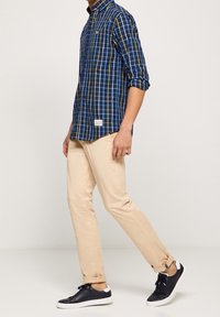 Modrá a žlutá kostkovaná košile s límcem, zkombinovaná s béžovými slim kalhotami a černými teniskami s bílou podrážkou. Košile má štítek na lemování.