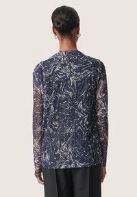 Bluză din material transparent, cu mânecă lungă, de culoare bleumarin, având un model abstract de valuri albe. Prezintă un decolteu rotund și o croială lejeră, fiind combinată cu pantaloni închisi la culoare.