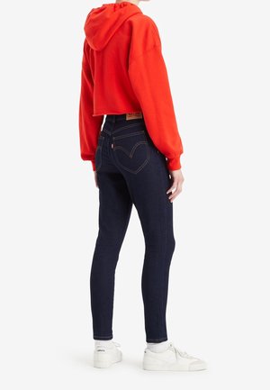 Person trägt dunkelblaue, hoch taillierte Jeans und einen kurzen roten Hoodie, steht seitlich mit weißen Sneakers und weißen Socken.