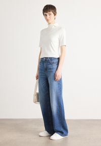 Hvit kortermet høyhalset skjorte parret med høytlivde vide blå jeans og hvite sneakers, holder en liten beige skulderveske.