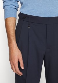 Main gauche reposant à côté d'un pantalon plissé bleu marine avec un bouton de réglage latéral et un anneau métallique, porté avec un pull en maille bleu clair.