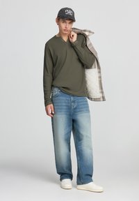 Maglietta a maniche lunghe verde oliva, jeans larghi azzurri, sneakers bianche, cappellino grigio con testo "New Jersey East Coast" e giacca a scacchi.