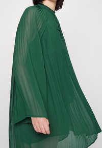 Zielona plisowana bluzka wykonana z przezroczystego materiału z wysokim dekoltem. Luźny krój, długie rękawy i miękki opad u dołu.