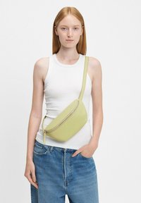 Limonkowa torebka noszona na przekór w gładkiej skórze, o zakrzywionym kształcie, z górnym zamkiem błyskawicznym i cienkim, regulowanym paskiem. Noszona na białym topie.