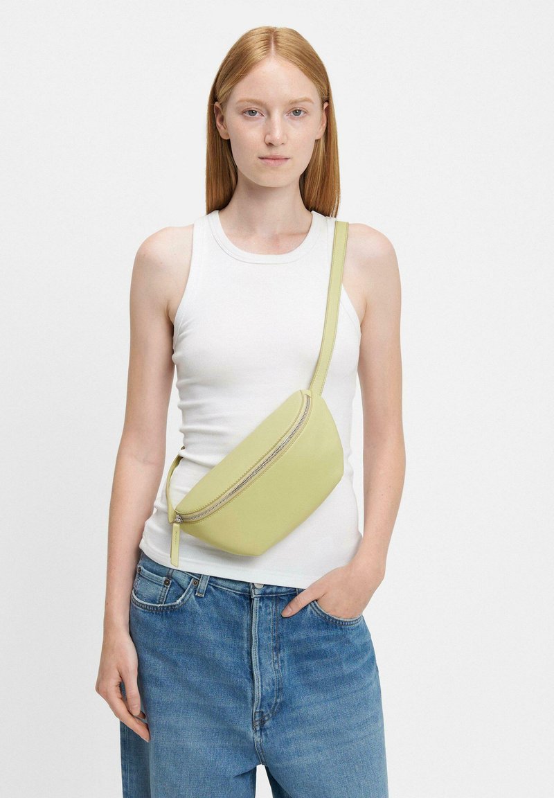 Limonkowa torebka noszona na przekór w gładkiej skórze, o zakrzywionym kształcie, z górnym zamkiem błyskawicznym i cienkim, regulowanym paskiem. Noszona na białym topie.