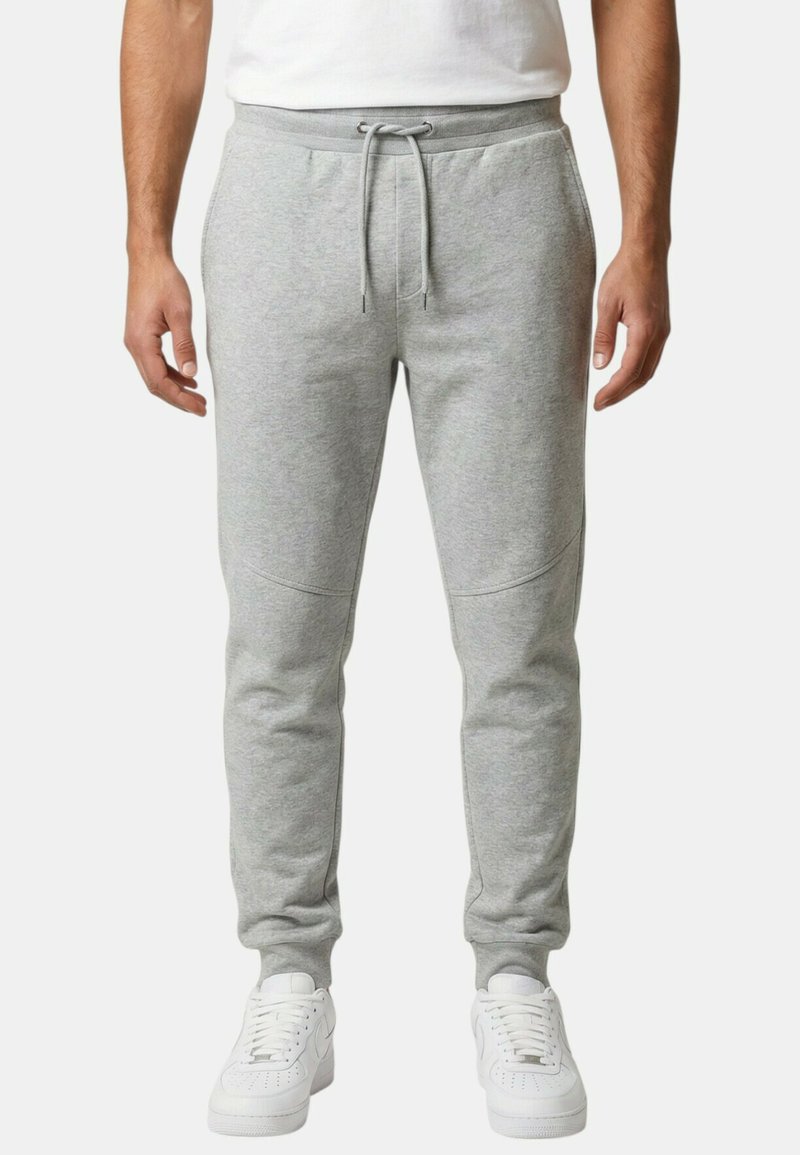 Homme portant un pantalon de jogging gris clair avec cordon de serrage et des baskets blanches, debout devant un fond uni.