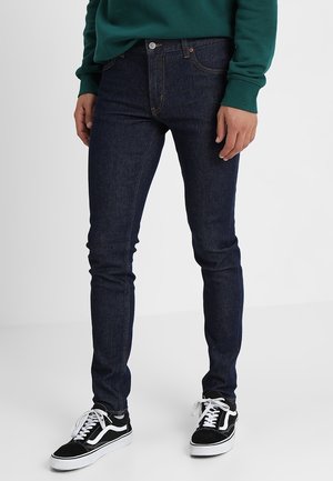 Smala mörka denimjeans med en slät textur. Har en knappstängning och fem fickor. Kombineras med svarta och vita sneakers.