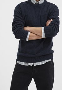 Mörkblå stickad tröja med ribbade ärmslut och nederkant, lager på lager över en grå och vit randig skjorta. Outfiten har en avslappnad passform.