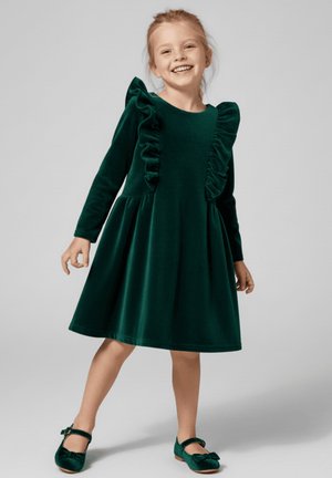 Zielona aksamitna sukienka z długimi rękawami, z falbankami z przodu i rozkloszowanym dołem. Zestawiona z pasującymi zielonymi butami.