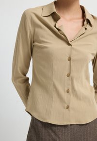 Blusa beige a maniche lunghe con colletto, apertura frontale con bottoni e tessuto testurizzato; presenta sottili linee orizzontali e un orlo curvo.
