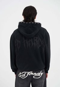 Sudadera negra con un gran logo negro de "Ed Hardy" en la parte posterior, con ojales metálicos en la capucha. Combinada con jeans oscuros que exhiben bordados en blanco de "Ed Hardy".