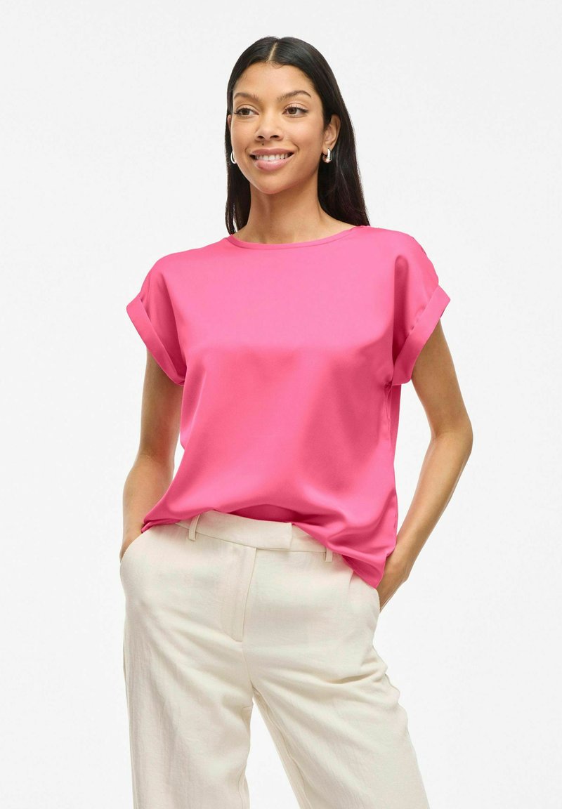 Top rosa a maniche corte con scollatura rotonda, texture liscia e orli delle maniche arrotolati, abbinato a pantaloni beige chiaro.