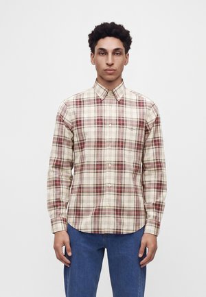 Junger Mann mit kurzen lockigen Haaren, der ein beiges und rotes kariertes Hemd mit Knöpfen und blaue Jeans trägt und vor einem weißen Hintergrund steht.