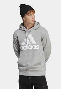 Felpa grigia con logo bianco Adidas sul petto, cappuccio con coulisse, tasca frontale e polsini a costine. Indossata con un berretto di lana nero.